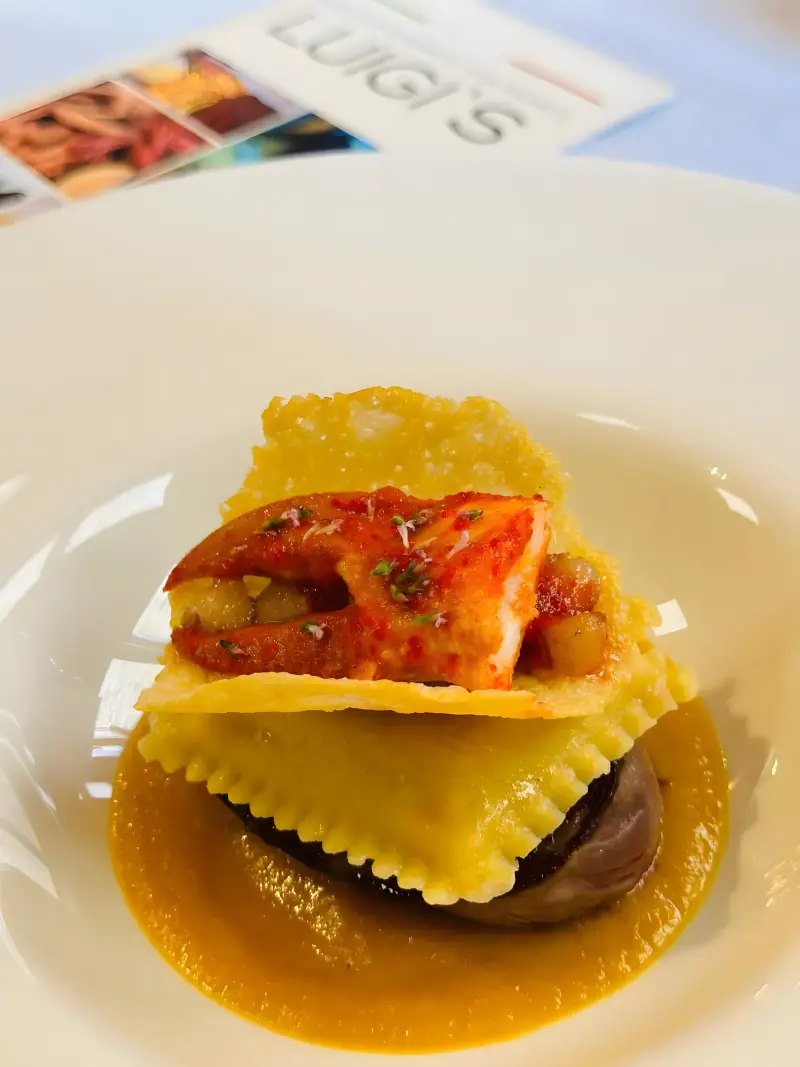 Ravioli on a golden sauce, topped with a parmesan crisp and a crab claw, with a Luigi’s menu behind the plate