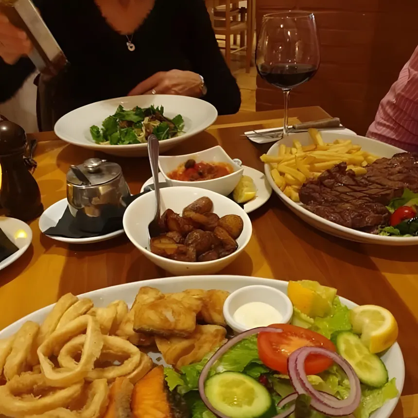 A table of fried seafood, grilled fish, salad, roast potatoes, steak and a glass of red wine.