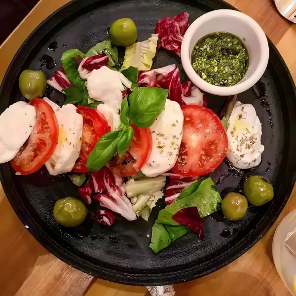 Sliced tomato and mozzarella with olives, radicchio and a pot of pesto on a dark plate.