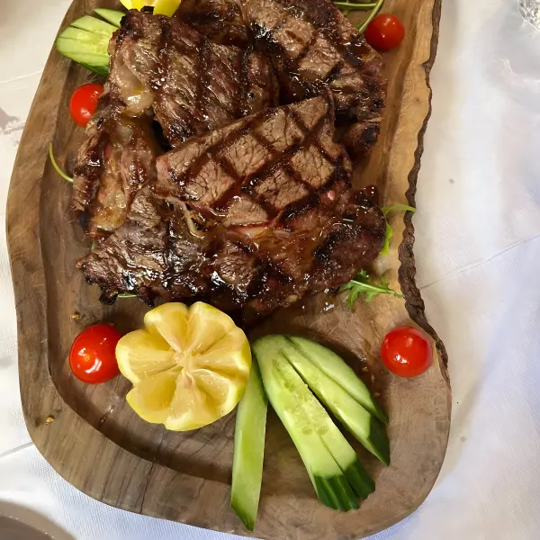A wooden platter of grilled steaks with lemon, cucumber, cherry tomatoes and greens.